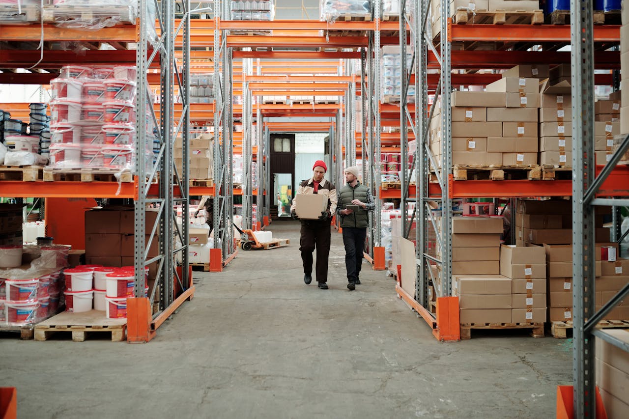 services-04 Two workers handle a package in a spacious warehouse surrounded by shelves stocked with boxes and products.